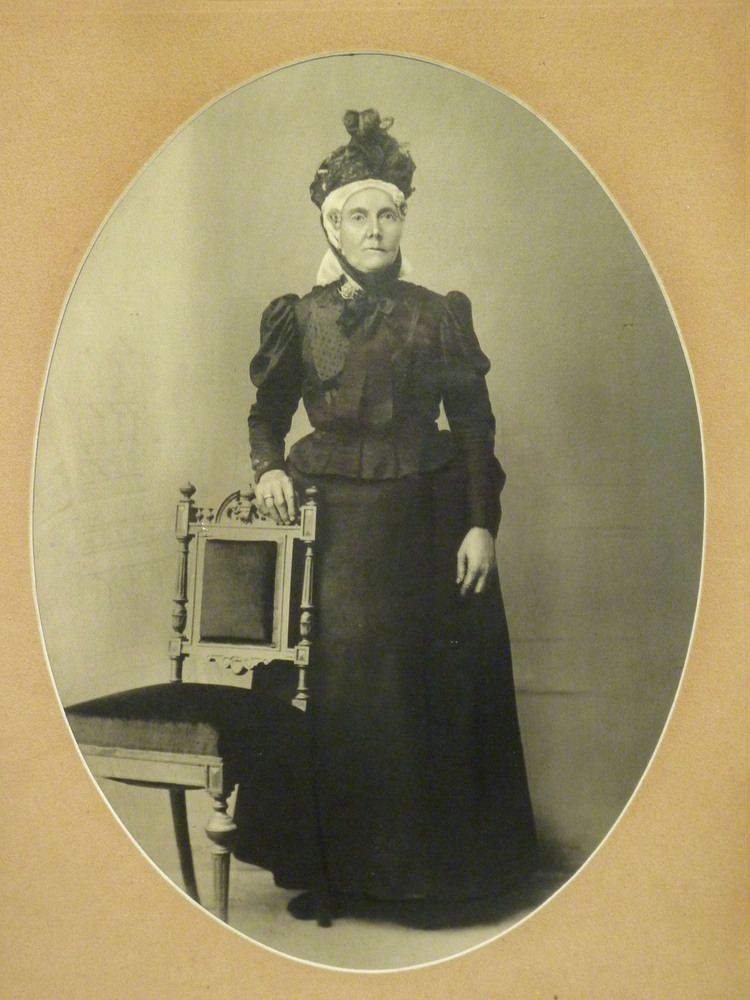 ovale foto van een vrouw met floddermuts en oorijzers en kapothoedje, staand naast een stoel, Geesje Drost-Braam: geb. 04-11-1833 te Wildervank, overleden 08-11-1911. Ze was toen ze op 10-02-1859 te Wildervank trouwde met Simon Drost dienstmeid. Ze was de moeder van Pietertje Lukkien-Drost en de grootmoeder van Simon Harm Lukkien en Hendrik Lukkien. Foto genomen omstreeks 1895-1900 door een fotograaf te Haren (Dld).