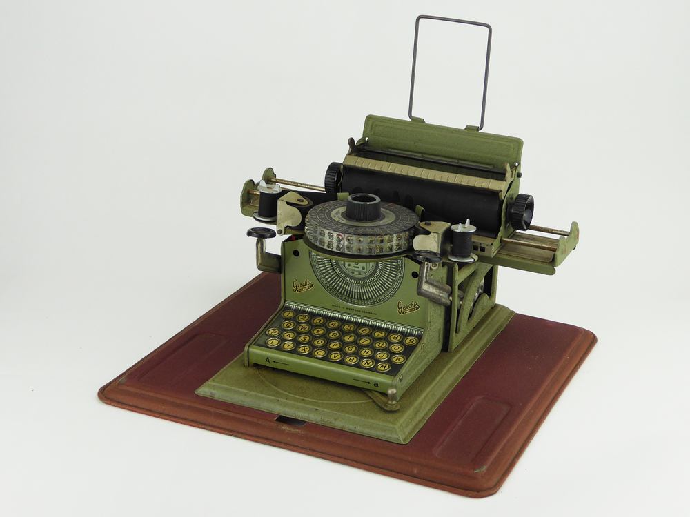 kleine groene metalen typemachine op bruine onderplaat, met bruine blikken opberghoes of koffer, in het midden draaibare metalen kop met dubbele letters, aan onderkant schuif voor kleine letters en hoofdletters, aan linkerkant knop voor doorspoelen lint, aan rechterkant knop voor afdrukken, aan voorkant inscriptie Geschir Patent made in western germany, aan voorkant beschermkoffer ingeslagen junior