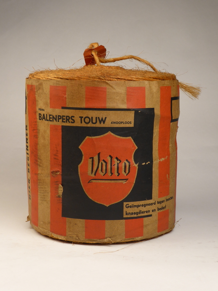 rol Balenperstouw, diameter 24,5 cm, hoogte 25,5 cm, met rood bruin geblokte wikkel met zwarte opdruk fabrikanten touwfabriek de volharding NV oude pekela , keerzijde Volto (Volharding Touwfabriek).