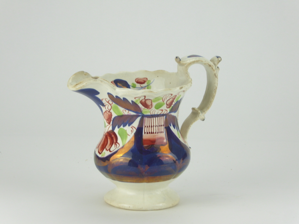 Witte aardewerken Engelse roomkan met tweeledige voluutoor, onder aanhechting oor een knobbel, gelijkende een blad, staand op een standvoet, met lange smalle schenklip, rondom kobaltblauwe font met roze lustre accenten en bruin paarse bloem en groene bladdecoraties, gelijkend op het Nederlandse boerenbontmotief, aan weerszijden wit vlak met bovenin koperkleurig traliemotief aan bovenzijde en bruin vlak eronder, tevens aan de binnenkant gedecoreerd met bloemmotieven. Patroon Gaudy Welch Stijl, geen merkjes op onderkant, ondanks hun naam gewoon in Engeland gefabriceerd, deed Imari porselein na, vervaardigd tussen 1820 – 1860.