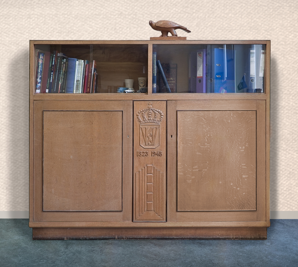 eikenhouten kast, bovenkant twee glazen schuifpanelen, onderkant 2 kasten met middenstuk met daarop gebeiteld een kroon met daaronder K & J W 1823 - 1948, gemaakt voor het 125 jarig jubileum van houtzagerij K & J Wilkens. Ze kregen toen het preadicaat Koninklijke.