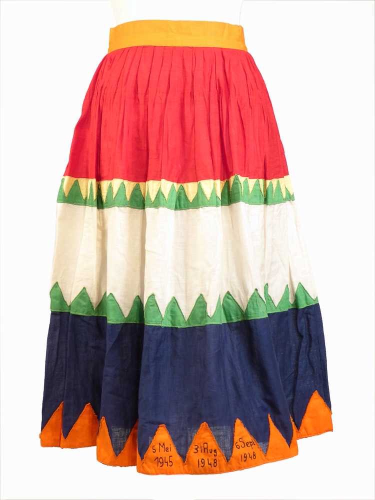 rood, wit blauwe katoenen rok met tussen de kleuren groen gele driehoeken, aan onderkant op oranje driehoeken zwarte belettering 5 mei 1945 31 aug 1948 6 sept 1948. Binnenin zit een stempel: B.J.Abeling Oude Pekela telefoon no 72. De kleuren staan symboliek voor : Oranje - huis oranje, rood wit blauw - Nederland, geel groen - vlag van Oude Pekela. Gebruikt door schenker G.M. Abeling. Geziena Maria Anna Abeling werd 4 september 1922 te Oude Pekela geboren als dochter van Bernhardus Johannes Josephus Abeling en Catharina Gesina Hensen. Haar ouders hadden een manufacturenzaak aan de Hendrik Westerstraat 471 te Oude Pekela met de naam "Hebo". De rok werd gedragen op bevrijdingsdag 5 mei 1945, op 31 augustus 1948 bij het 50 jarig jubileum van Koningin Wilhelmina en op 6 september 1948, bij de inhuldiging van Koningin Juliana.