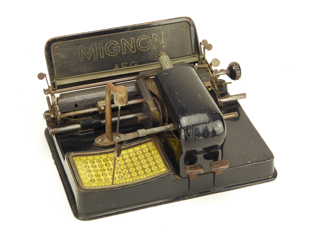 rechthoekige metalen constructie met geel toetsenbord  / letterbord, en neerwaartse naald, vastgehouden door aan rechterkant aanwezige metalen constructie, op achterplaat merk Mignon AEG