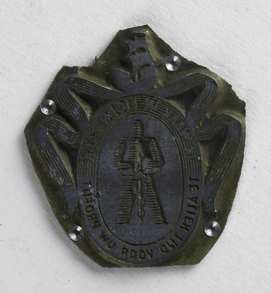 ietwat ovale non ferro metalen cliche met rechthoekige bovenstukje, met afbeelding ovaal met hierin harnas die esculaapvormige zwaard vasthoud, boven rondom banier met op de top een zeilschip, rondschrift om ovaal ik waak in eigen zaak te allen tijd voor uw profijt ( wapenspreuk van de Stichting Reclame Middenstand), afkomstig van kantoorinstallaties Bv Schuur te Veendam