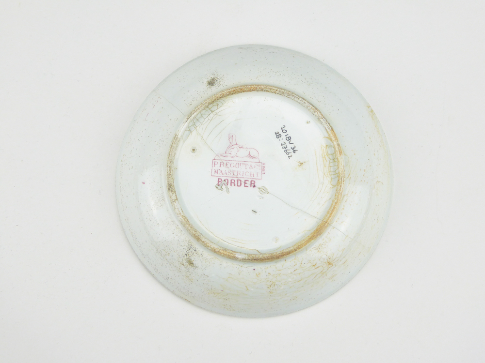 aardewerken schotel van het merk Petrus Regout & Co, Maastricht, decornaam Border, diameter 13,3 cm, op binnenkant rode decoratie van bloemen en wolfsangel, gemerkt met beeldmerk 71 A van Petrus Regout, blindmerk 4, opgedoken door British Sub Aqua Club, Branch 501, ook wel de 501 Divers uit Folkstone, uit het wrak van de op 19 november 1887 na een aanvaring met het stoomschip Rosa Mary gezonken stoomschip W.A. Scholten, via hun woordvoerder Glyn Jones geschonken aan het Veenkoloniaal Museum.