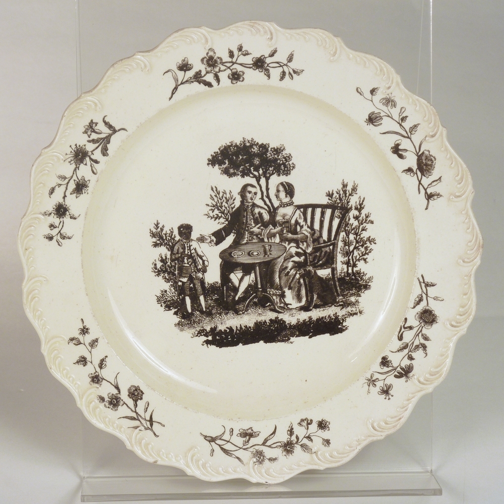 aardewerken bord met geschulpte rand met zwarte afbeelding drinkend paar in tuin met bediende en bloemmotief met rankwerk op rand