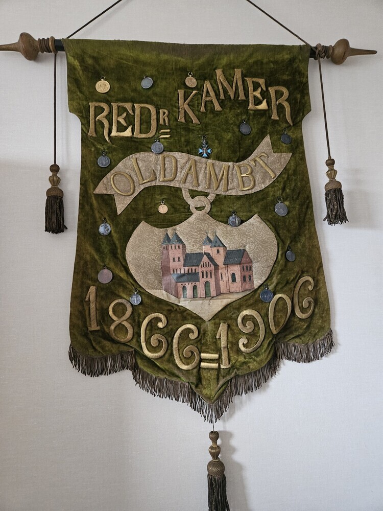 groen velours wapenschild met goudkleurige geborduurde belettering Red.R. Kamer Oldambt 1866 - 1906, met in het midden in cartouche een kerk met torens, de Viertorenkerk te Midwolde, hangende aan een stok met puntige zijkanten en kwasten, aan de onderkant ook een kwast. Het zijn houten vormpjes omwonden door metaaldraad met een katoenen kern. Het geheel is behangen met 15 medailles.