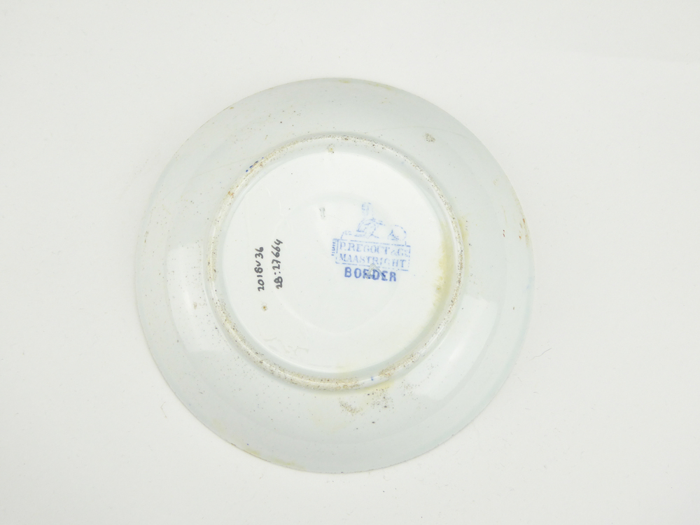 aardewerken schotel van het merk Petrus Regout & Co, Maastricht, decornaam Border, diameter 13,5 cm, op binnenkant blauwe decoratie van bloemen en wolfsangel, gemerkt met beeldmerk 71 A van Petrus Regout, blindmerk 4, opgedoken door British Sub Aqua Club, Branch 501, ook wel de 501 Divers uit Folkstone, uit het wrak van de op 19 november 1887 na een aanvaring met het stoomschip Rosa Mary gezonken stoomschip W.A. Scholten, via hun woordvoerder Glyn Jones geschonken aan het Veenkoloniaal Museum.