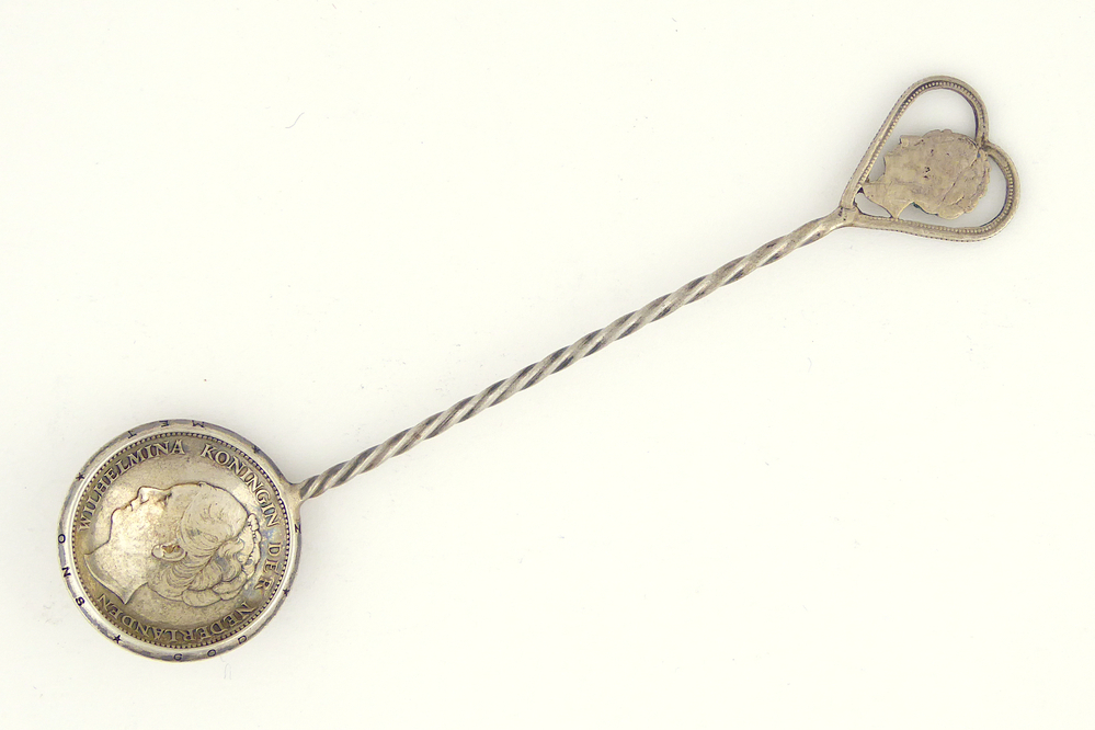 zilveren lepel met ronde bolle bak bestaande uit een zilveren gulden uit 1940 en hartvormige constructie waarin een uitgezaagde kwartje van Wilhelmina uit 1941 aan uiteinde steel. Tot het jaar 1941 werden er zilveren munten geslagen met de beeltenis van Koningin Wilhelmina. In 1942 werden echter de zinken munten geïntroduceerd. Enerzijds omdat het zilver gebruikt kon worden ten bate van de Duitse oorlogsindustrie. Anderzijds omdat het de Duitsers stoorde dat de beeltenis van Koningin Wilhelmina op de munten te vinden was. Vervolgens werden er van de zilveren munten sieraden gemaakt, waaronder broches, armbanden, speldjes, lepeltjes en hangers. Door het te dragen kon je aangeven dat je loyaal was naar het Nederlandse koningshuis. Dit was overigens niet zonder gevaar!