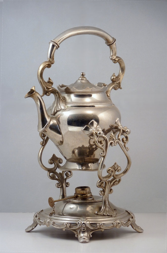 bouilloire. Een bouilloire of waterketel op bijpassend komfoor, die diende om in de huiskamer het theewater aan de kook te houden.  Hiervoor stond de ketel op een komfoor die was gevuld met gloeiende kooltjes of zoals bij deze een petroleumbrander. Stamt origineel uit eind 17 e eeuw, toen het theedrinken in wijdere kring ingang vond. Thee was een duur artikel dat werd getrokken in een klein potje, dat regelmatig met heet water werd bijgevuld.