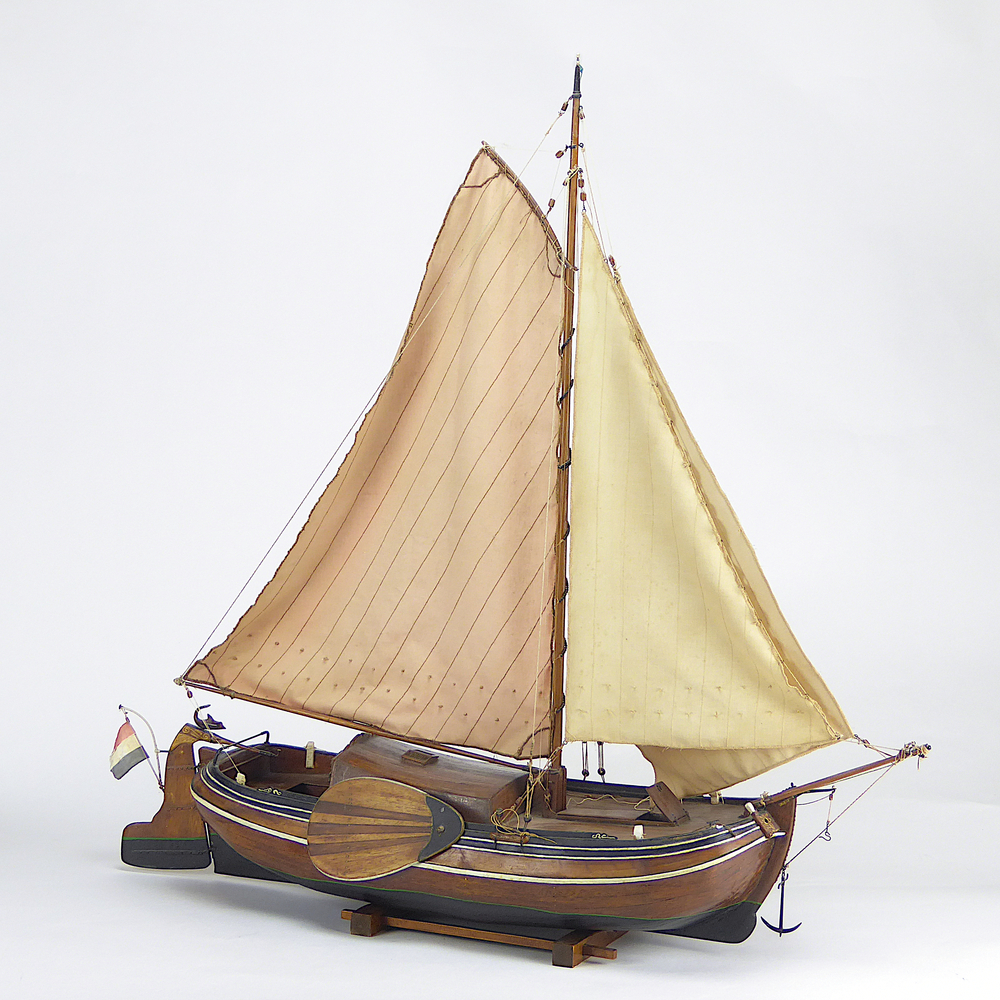 Houten scheepsmodel van een platbodem, met koperen accenten en rijkelijk versierd met houtsnijwerk, achterop roer een roerklik in de vorm van een vis / dolfijn en rood wit blauwe vlag aan roer, blauwe pijl in vlaggenmast top, staande op een houten onderstel, vervaardigd door Günther Zochorek, oud zeeman . Het schip heeft hij in 1979 gebouwd te Veendam.