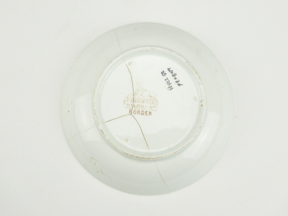 aardewerken schotel van het merk Petrus Regout & Co, Maastricht, decornaam Border, diameter 13,4 cm, op binnenkant bruine decoratie van bloemen en wolfsangel, gemerkt met beeldmerk 71 A van Petrus Regout, opgedoken door British Sub Aqua Club, Branch 501, ook wel de 501 Divers uit Folkstone, uit het wrak van de op 19 november 1887 na een aanvaring met het stoomschip Rosa Mary gezonken stoomschip W.A. Scholten, via hun woordvoerder Glyn Jones geschonken aan het Veenkoloniaal Museum.