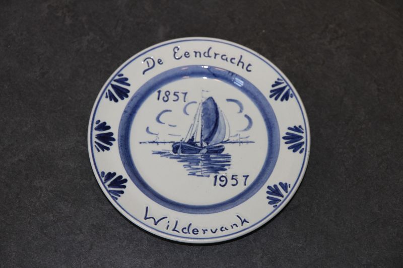 ronde gedenkbord met in blauw op voorkant inscriptie : de eendracht 1857 - 1957 wildervank (verzekeringsmaatschappij voor binnenschepen) Op voorkant afbeelding zeilschip, op achterkant inscriptie Delfts Blauw (Logo Ram) Ram Made in Holland en initialen ceramist WD, ontwerp Nico Bulder